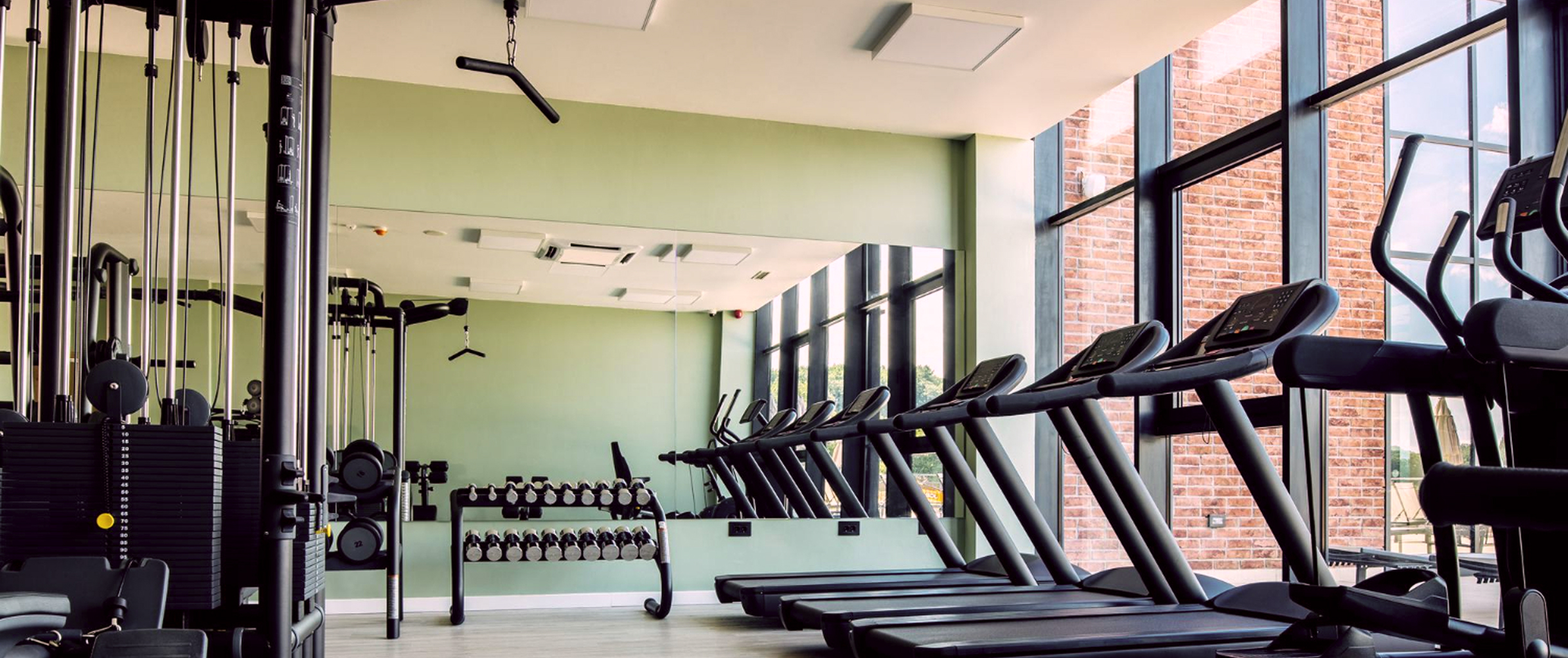 Тренажёрный зал, справа изображены беговые дорожки около окон, посередине стойка
    гантелями, слева кроссовер для упражнений с весом